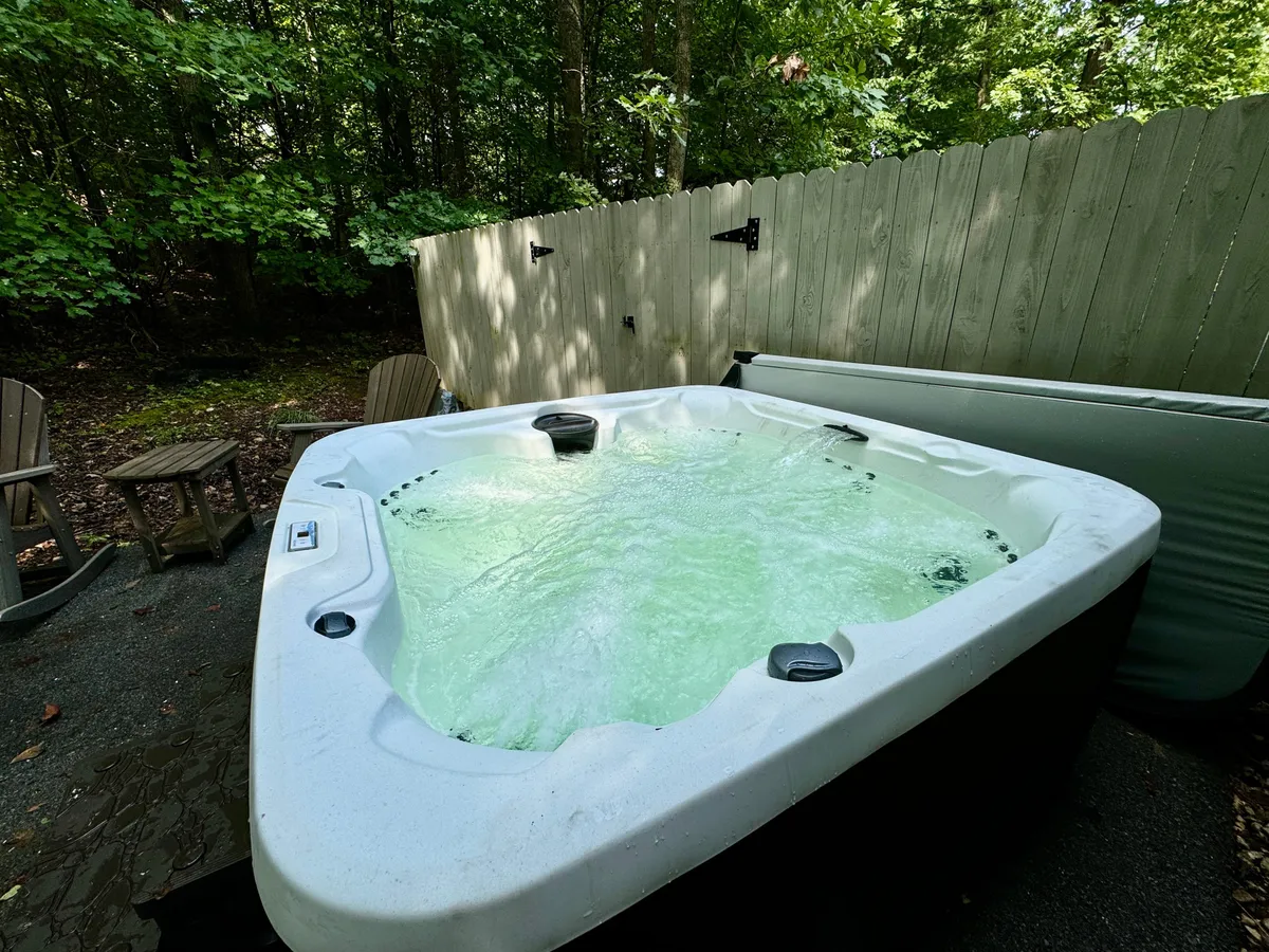 An exterior view featuring a private hot tub set amidst lush foliage, with wooden chairs nearby that invite relaxation and socializing. The natural setting offers both privacy and a sense of immersion in nature, creating an ideal spot for winding down and stargazing in the evening.