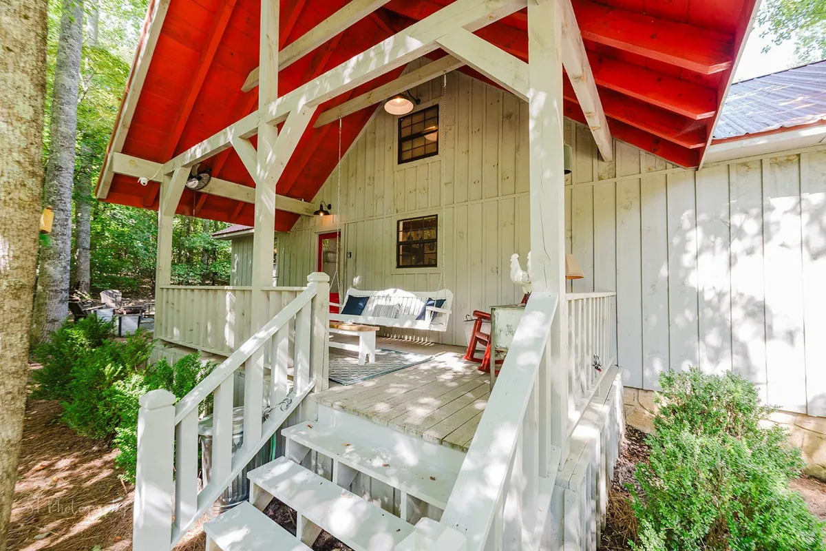 An exterior perspective of the cabin with its distinctive red roof and wooden porch, featuring swing seating, rocking chairs, and surrounding greenery. The scene is both welcoming and tranquil, suggesting quiet mornings and lively gatherings with family.