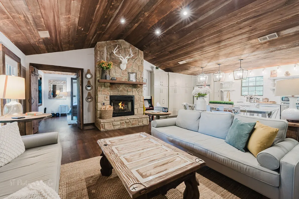 This interior shot prominently features the stone fireplace, wooden beams, comfortable sofas, and a rustic coffee table, with the kitchen area visible in the background. The arrangement suggests an open-plan layout perfect for socializing and relaxing.