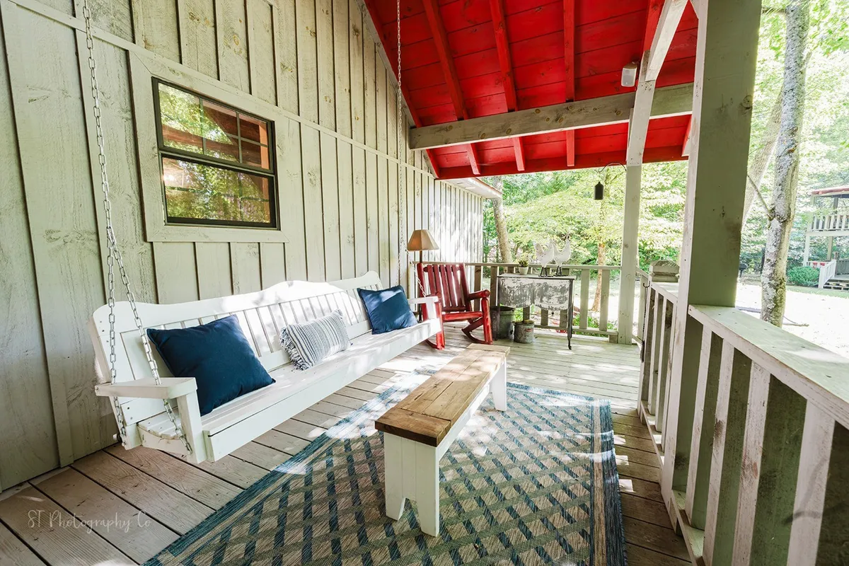 A welcoming outdoor scene showing a white swing bench, red chairs, and a wooden bench on a cozy rug. The arrangement is nestled among trees, suggesting a peaceful spot for relaxing, reading, or enjoying nature with loved ones.