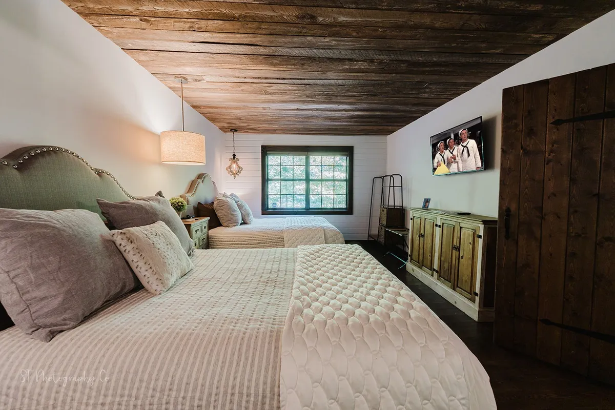 A spacious bedroom with two cozy queen beds, a large window offering woodland views, a wooden ceiling, a wooden cabinet, and a TV screen, creating a warm, inviting retreat for families or friends.