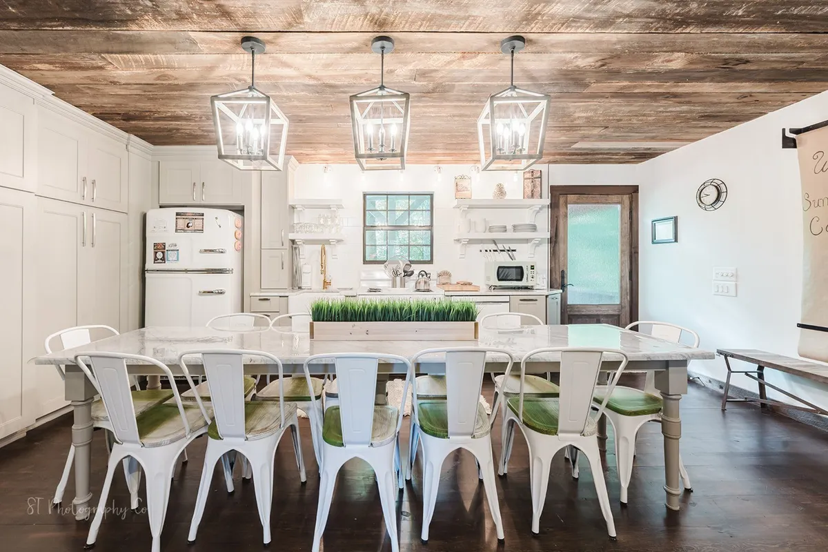 This image displays a large dining table set beneath hanging lantern-style lights, with a lush greenery centerpiece and white cabinetry visible in the background. The arrangement of seating suggests ample space for large gatherings, while the ambient lighting and homely décor evoke a sense of warmth and hospitality.