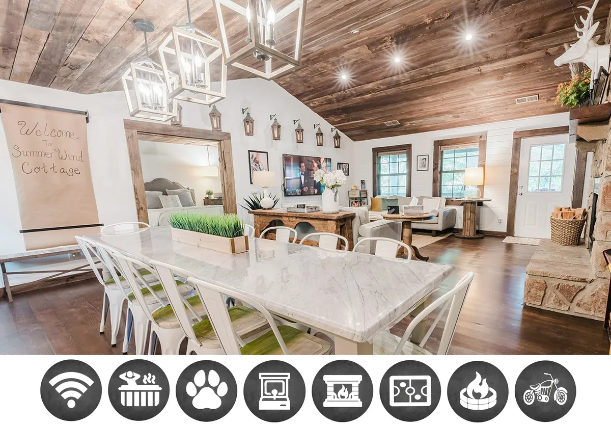 The image captures the warm, inviting interior of the Summer Wind cabin, featuring a spacious long dining table set for family gatherings, eye-catching hanging lanterns that cast a soft glow, a rustic stone fireplace as the cozy centerpiece, and a plush seating area designed for relaxation. The overall ambiance evokes comfort and togetherness, ideal for memorable moments.