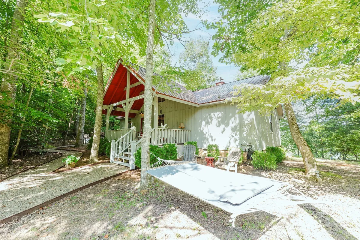 A peaceful backyard scene featuring a hammock strung between trees, the house with red accents, and a spacious porch. The area is surrounded by greenery, perfect for both quiet moments and family barbecues.