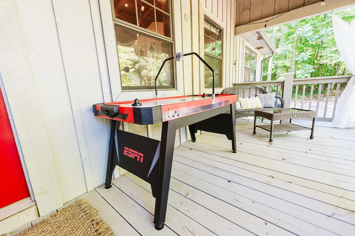 An exterior shot of a covered porch area with an air hockey table, outdoor seating, and views of natural greenery. The space is inviting for friendly competitions or relaxing with snacks and drinks.