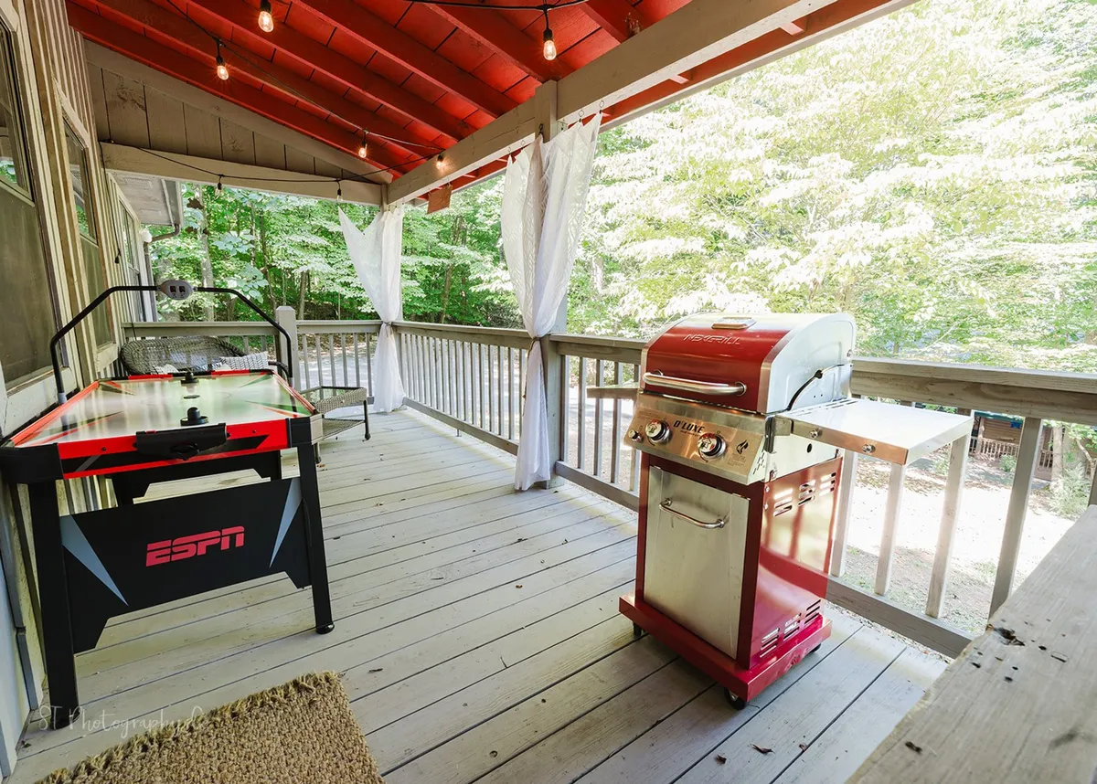 This exterior shot showcases a covered patio with an air hockey table, outdoor grill, decorative string lights, and comfortable seating, all set against a backdrop of natural greenery. The setup is playful, inviting, and perfect for both daytime activities and relaxed evenings.