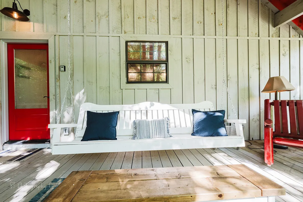 A cozy outdoor nook featuring a white swing bench with blue pillows, a red front door, and a wooden coffee table, set on a charming porch. The scene is perfect for morning coffee, reading, or quiet conversations, with a relaxed and welcoming vibe.