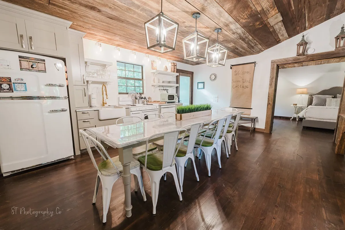This interior scene focuses on a spacious dining table adjacent to a kitchen counter, with inviting lighting fixtures and decorative wall art. The open layout connects the kitchen and dining spaces, facilitating both cooking and socializing.