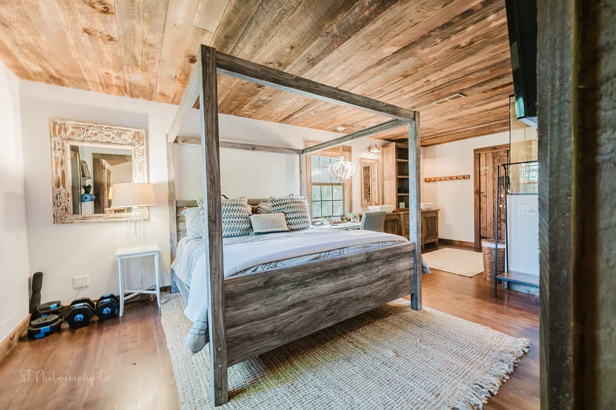A serene primary bedroom featuring a four-poster bed with cozy linens, a decorative mirror, textured rugs, and a wooden ceiling. Natural light filters softly through the window, and the tranquil setting invites relaxation and rest.