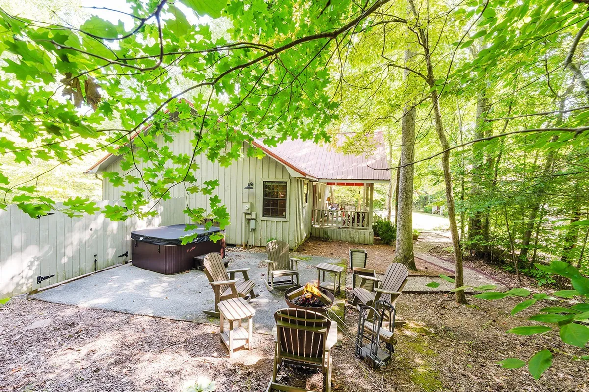 This image shows a cozy outdoor area with a hot tub, fire pit surrounded by wooden chairs, the house exterior, and lush trees. The setting invites both relaxation in the hot tub and social time around the fire pit, with plenty of natural beauty in the background.