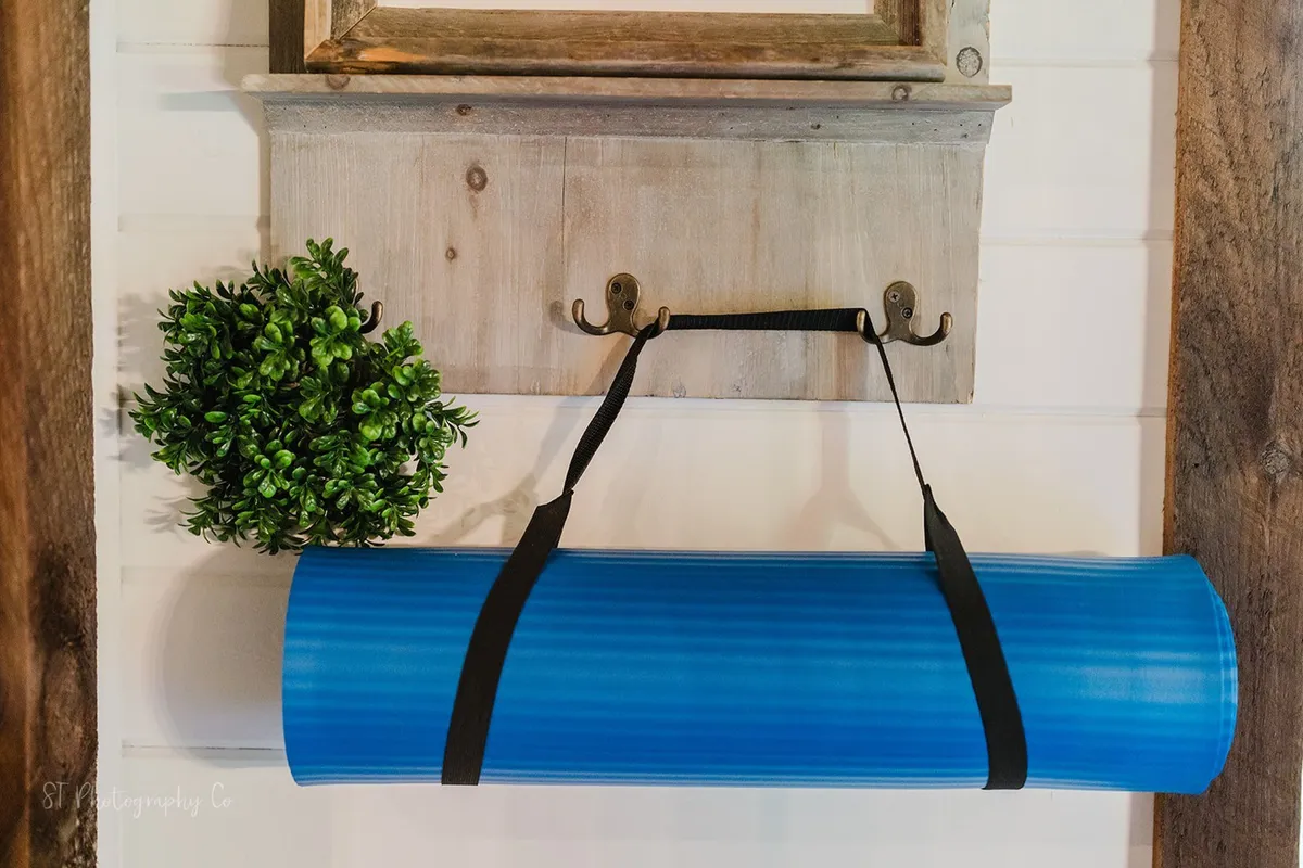 A quiet corner featuring a blue yoga mat, green plant, wooden hooks, and a framed picture, evoking a calming environment for yoga, stretching, or meditation.