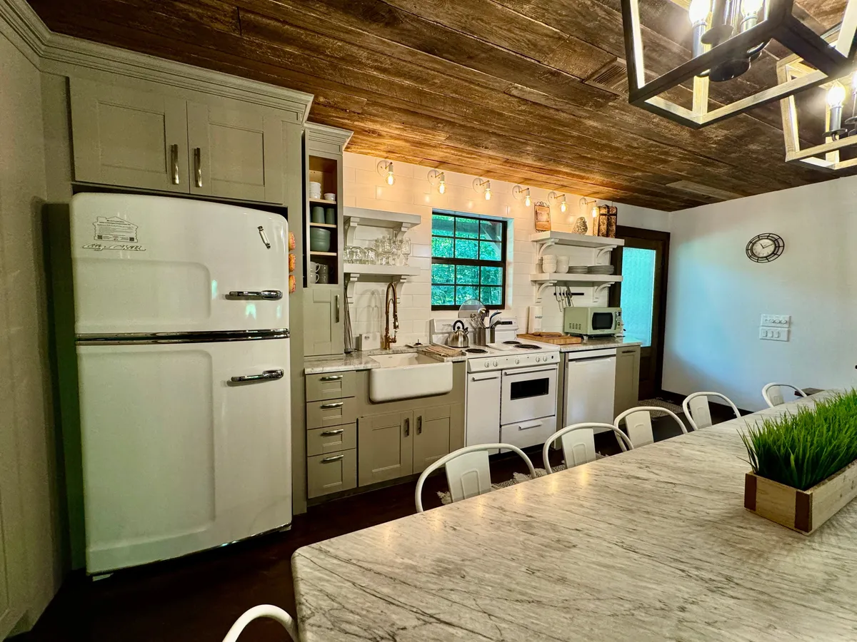 A bright kitchen scene featuring a white refrigerator, marble dining table, wooden ceiling, window above the sink, and visible kitchen appliances. The space looks well-equipped and practical, hinting at modern convenience and thoughtful touches.
