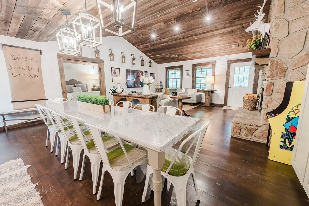 This angle shows a long dining table, stone fireplace, and adjacent couch area, all under soft light from chandeliers. The kitchen is visible in the background, framing a cohesive open-concept living and dining space.